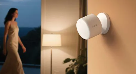 A white Aqara sensor is mounted on the wall in a warm room, while a woman walks past in the background.
