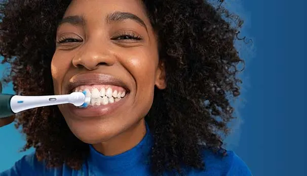 Een vrouw met volle krullen poetst lachend haar tanden met een elektrische tandenborstel, voor een effen blauwe achtergrond in close‑up.