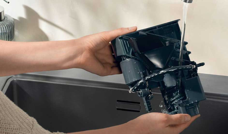 Een persoon reinigt het uitneembare zetgroep-onderdeel van een Siemens koffiemachine onder stromend water bij de gootsteen.