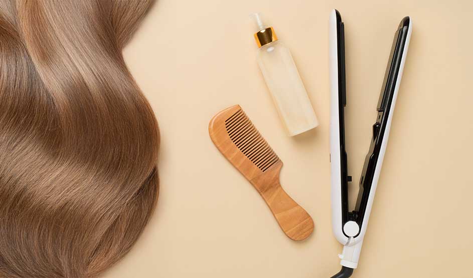 Wavy, glossy hair lies beside a comb, a spray bottle and a hair straightener on a soft beige surface.