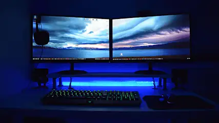 Twee computerschermen tonen een landschap onder een blauwe gloed, met een toetsenbord en muis op een bureau.