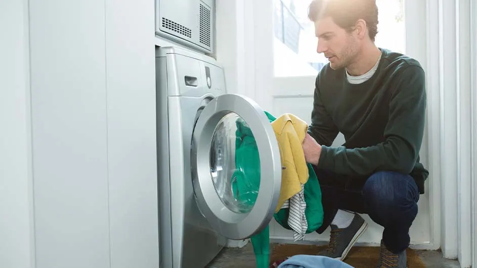 Een man hurkt voor een open wasmachine en haalt er gekleurde kleding uit. Naast de wasmachine staat een droger.