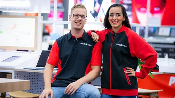 Twee mensen in rood-zwarte uniformen glimlachen in een winkel. Ze staan voor tafels met elektronische apparaten en schermen.