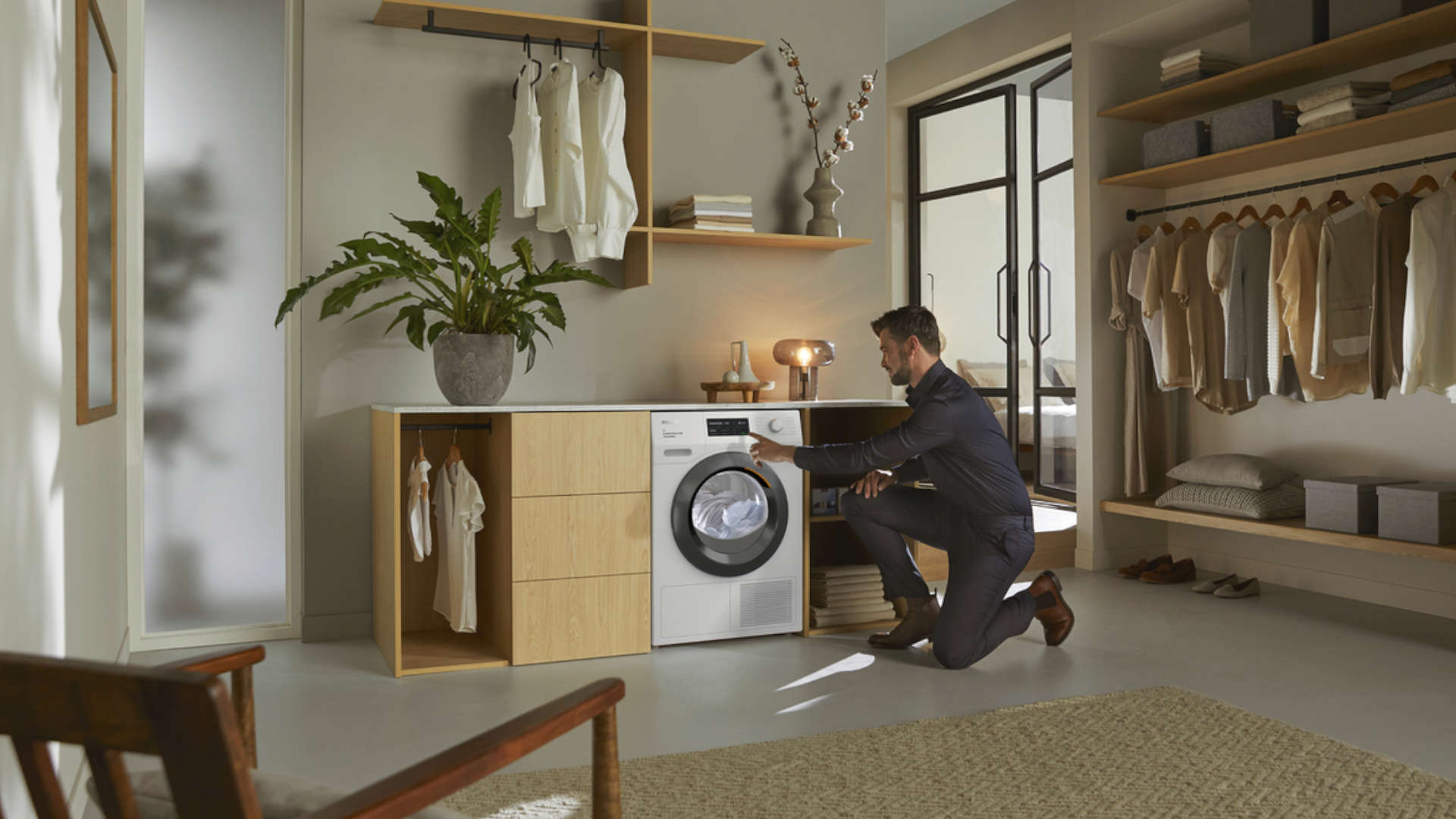 Een man knielt voor een wasmachine in een kamer met kasten en planken vol kleding.