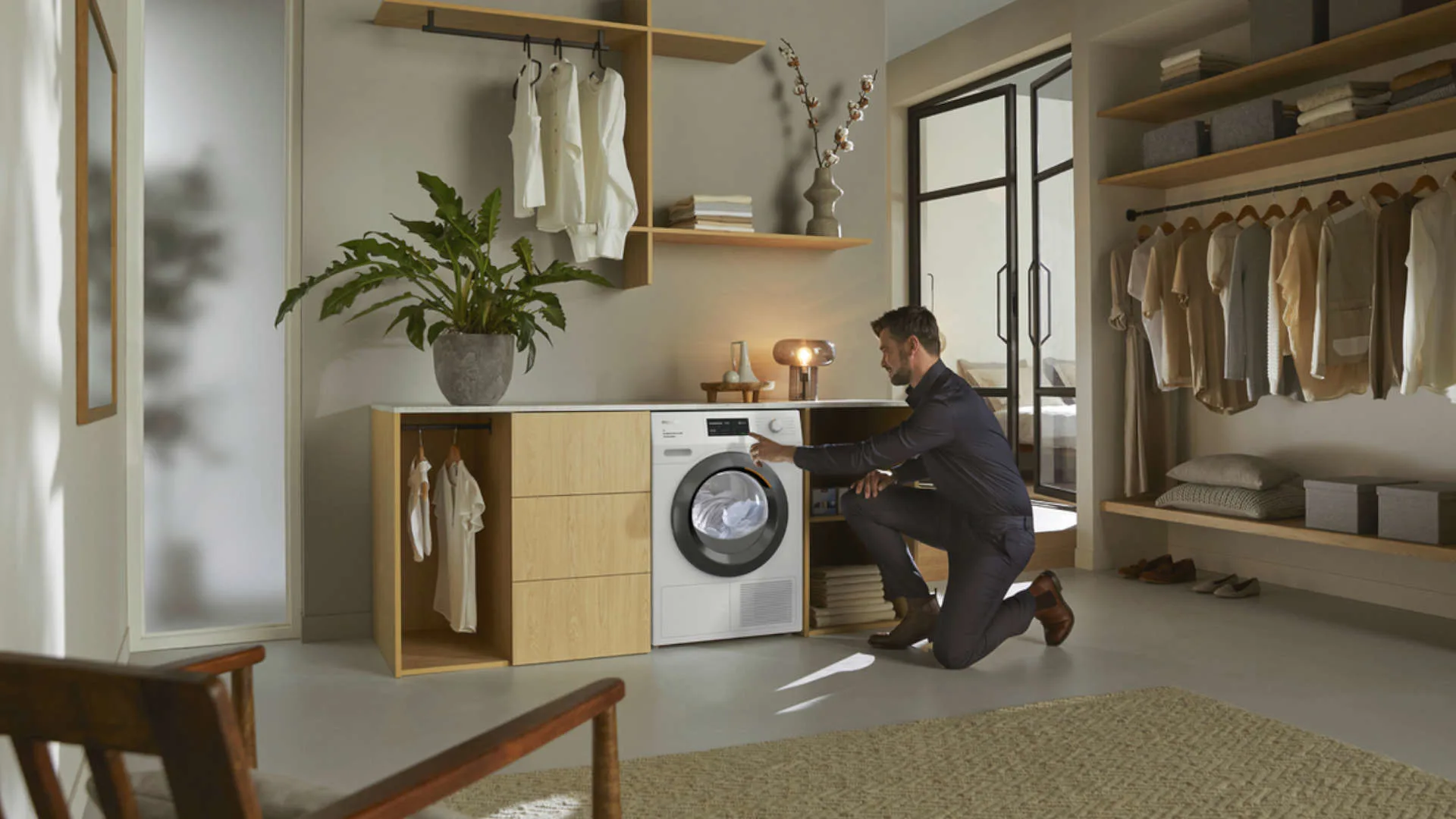 Een man knielt voor een wasmachine in een kamer met kasten en planken vol kleding.