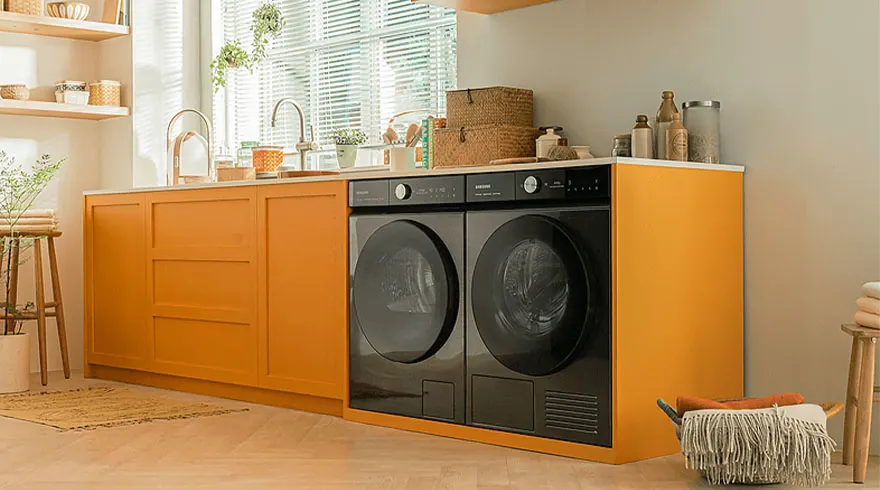 Waskamer met oranje kast, aanrecht met gootsteen, zwarte wasmachine en droger. Mand met deken en houten kruk.