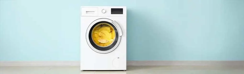 Een witte wasmachine staat tegen een blauwe muur op een witte vloer. De machine bevat gele was.
