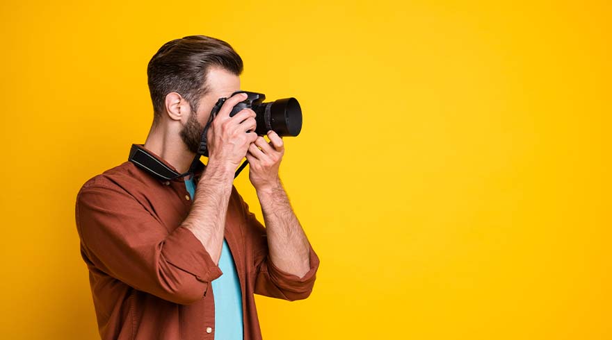 Een man met een camera tegen een gele achtergrond. Hij draagt een bruin overhemd en een blauw shirt.