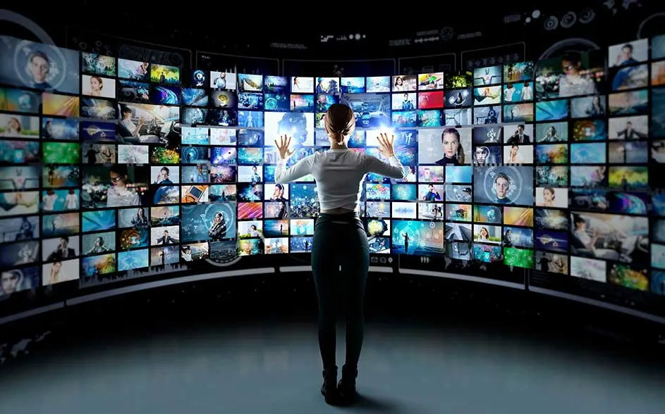 Een vrouw staat met opgeheven handen voor een gebogen muur vol schermen met foto’s en video’s; het lijkt op een futuristische mediacentrale.