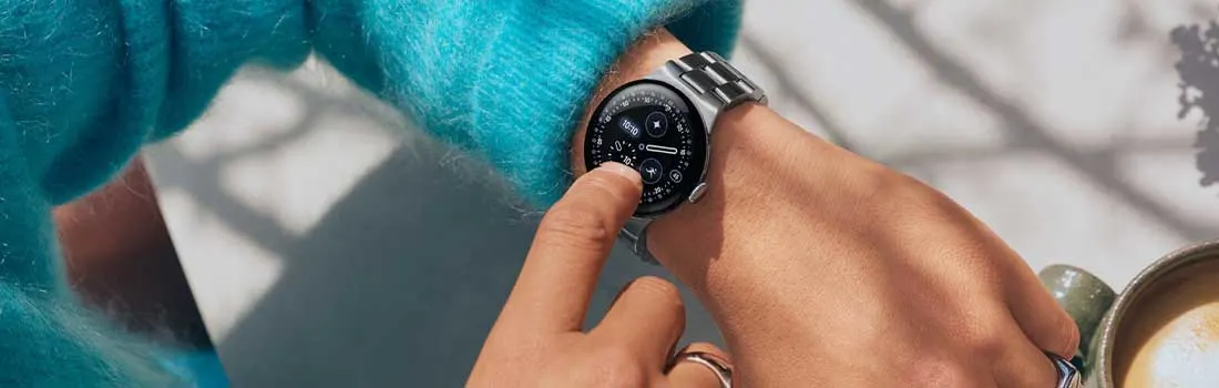 Een Google Pixel Watch 4 smartwatch met metalen band wordt bediend met aanraking op het scherm, naast een kop koffie.
