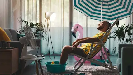 Man relaxes in a chair with a foot bath under a striped umbrella inside. A fan, lamp, and flamingo float are also visible.