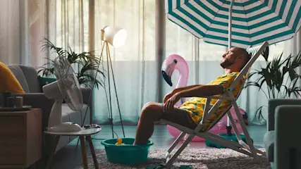 Een man ontspant in een strandstoel in een woonkamer met ventilator, flamingo, parasol en een teiltje met water.