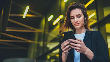 Een vrouw in een zwart jasje kijkt naar een zwarte telefoon in haar handen, met gele lichten op de achtergrond.