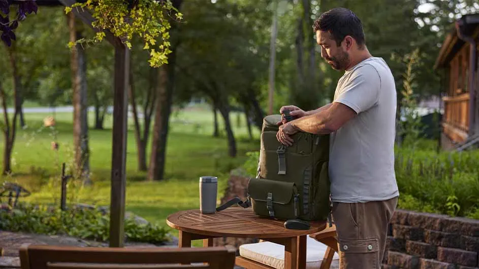 Man pakt een groene rugzak in op een houten tafel met een beker. Een tuin en een houten huis staan op de achtergrond.