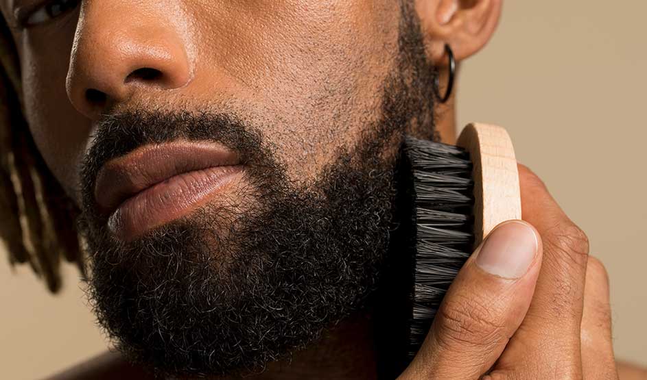Een close-up toont een man met volle baard die met een houten borstel zijn baard verzorgt; hij draagt een oorring.