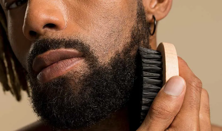 A close-up shows a man with a full beard grooming it with a wooden brush; he is wearing an earring.