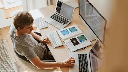 Een man zit aan een bureau met laptops, een toetsenbord, een computerscherm en een notitieboek met afbeeldingen.