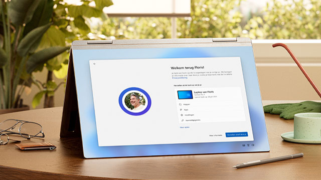 Laptop toont 'Welkom terug' scherm. Naastgelegen: bril, portemonnee, stylus, tuinhandschoenen en een kop. Raam en plant op achtergrond.
