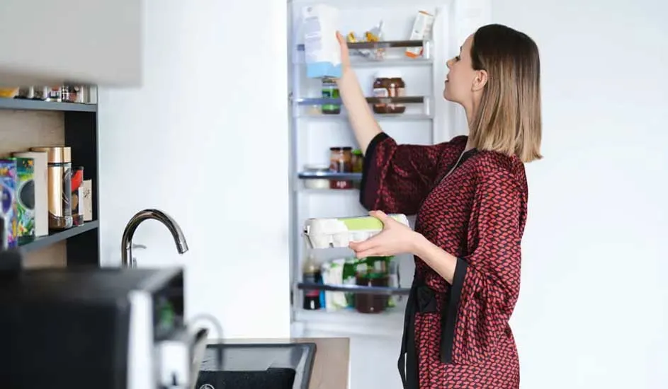 Een vrouw in een rode kamerjas pakt een pak melk uit de koelkast en houdt een doos eieren vast in een moderne keuken.