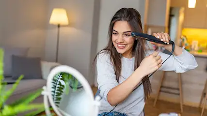 Een vrouw lacht en gebruikt een stijltang in een kamer met een spiegel en een lamp. Achtergrond toont een bank en keuken.