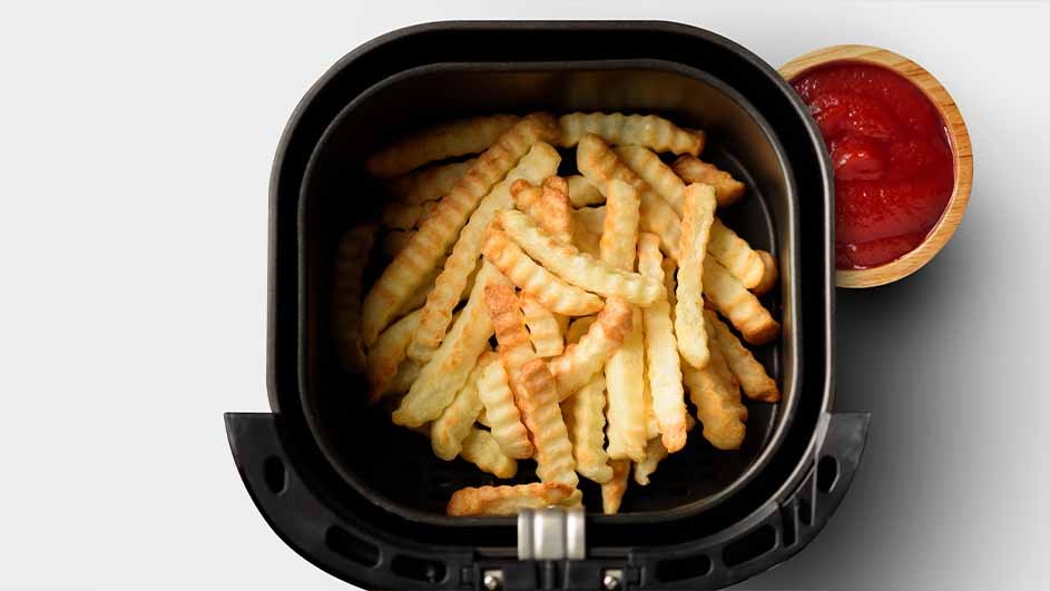 Crinkle-cut fries fill a black air fryer basket, with a small wooden bowl of red ketchup next to the fryer.