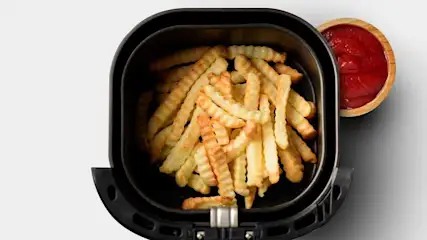 Crinkle-cut fries fill a black air fryer basket, with a small wooden bowl of red ketchup next to the fryer.