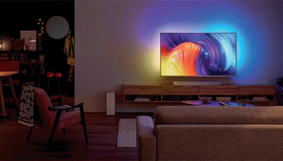 Woonkamer met tv aan de muur boven een houten kast. Een bank staat in de kamer en een stoel met een deken.