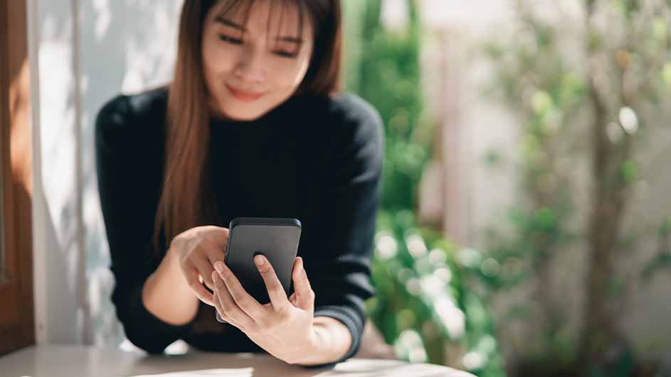 Vrouw in zwarte trui gebruikt een zwarte smartphone aan een tafel. Op de achtergrond zijn planten te zien.