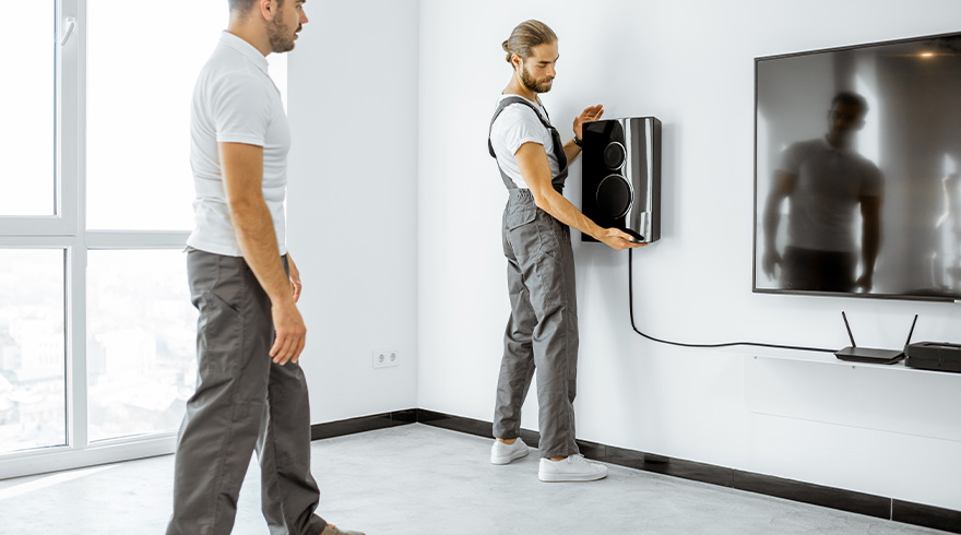 Twee mannen installeren een luidspreker naast een tv in een witte kamer.