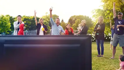 Groep mensen kijkt naar een tv buiten, sommigen juichen. Gras en bomen op de achtergrond.