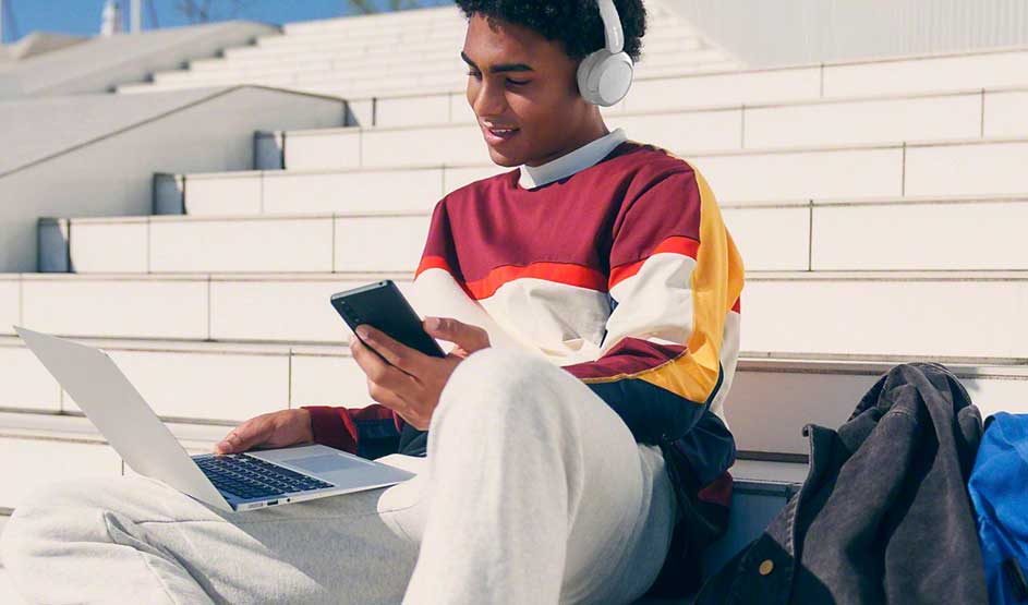 Iemand zit op traptreden buiten, draagt een witte Sony koptelefoon, gebruikt een laptop en houdt een smartphone vast, met een tas naast zich.