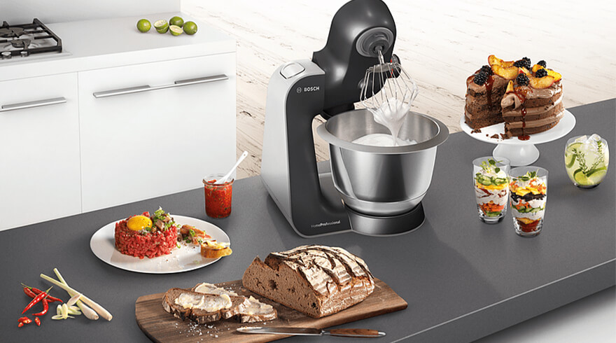 Een keukenopstelling met een mixer, taart, brood en gerechten op een grijze aanrecht.