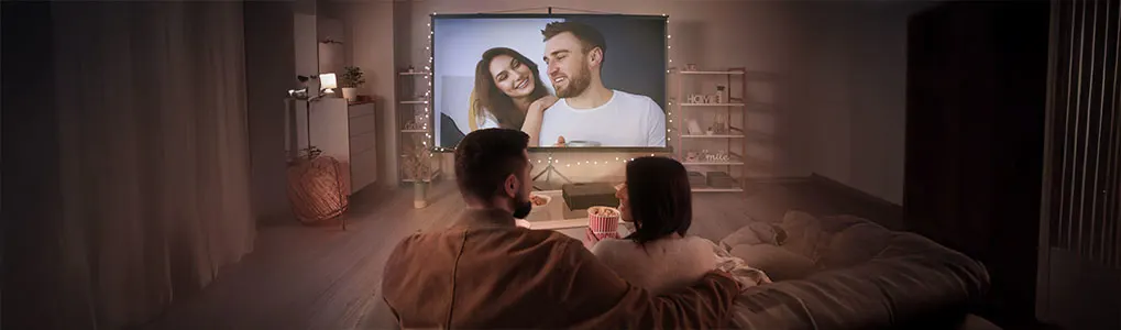 A couple watches a TV screen in bed; a woman and a man are on the screen. Popcorn and shelves are visible.