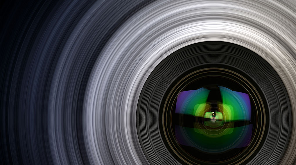 Close-up van een cameralens met reflecties van groen, blauw en paars. Grijze, concentrische lijnen vullen de achtergrond.