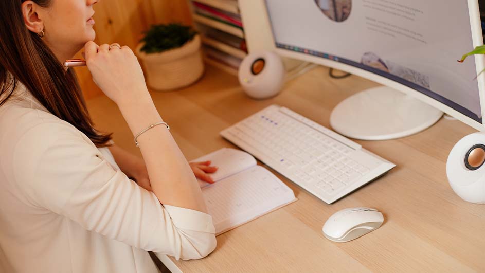 Een vrouw zit aan een bureau met een computer, toetsenbord, muis en een notitieboek.