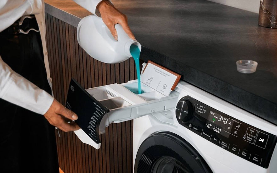 Een persoon giet blauw wasmiddel in de lade van een AEG wasmachine. Het display van de machine toont diverse instellingen, terwijl de omgeving een modern aanrechtblad en houten panelen laat zien.