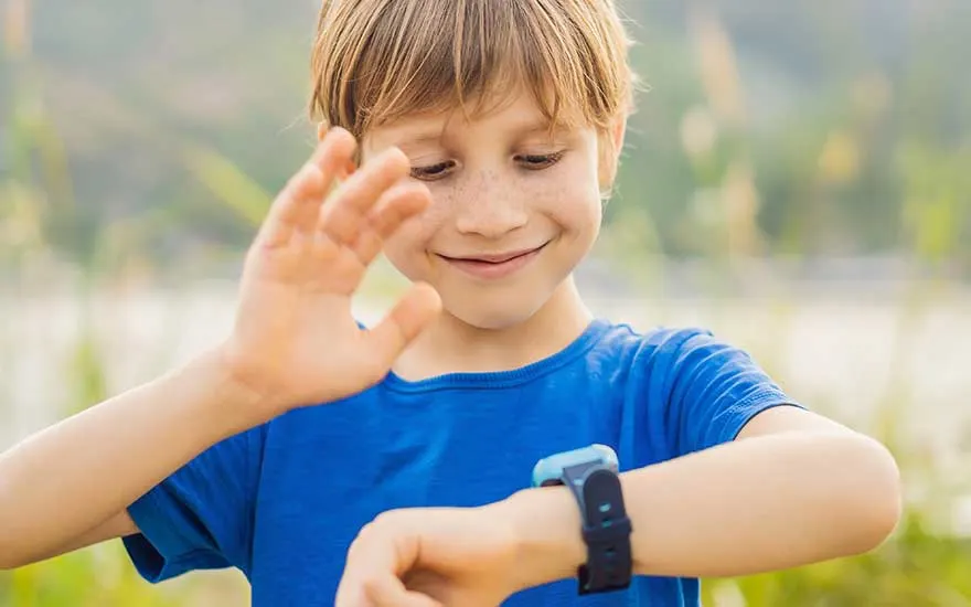 Een jongen met sproeten kijkt glimlachend naar zijn slimme horloge buiten, terwijl hij met één hand enthousiast naar het scherm zwaait.