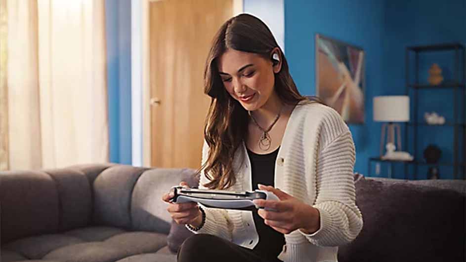 Vrouw zit op een grijze bank en speelt op een grijze spelcomputer met witte oortjes in, in een blauwe kamer.