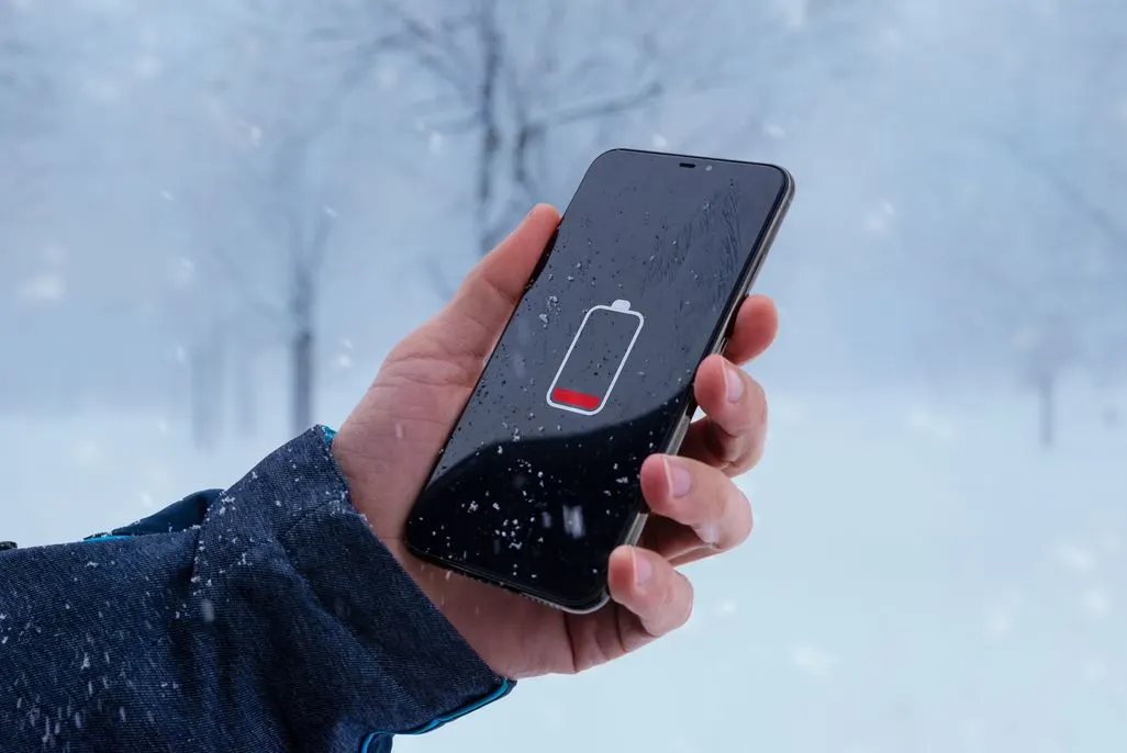 Hand in blauer Jacke hält schwarzes Smartphone mit leerem Akku-Symbol in verschneiter Winterlandschaft bei Schneefall.