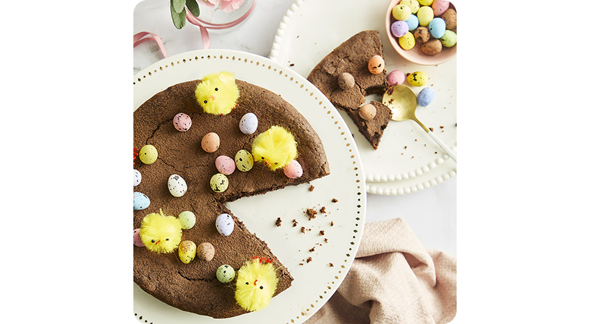Runder Schokokuchen mit herausgeschnittenem Stück, dekoriert mit bunten Schoko-Ostereiern und gelben Deko-Küken auf Tortenplatte, daneben weiterer Kuchenteller.