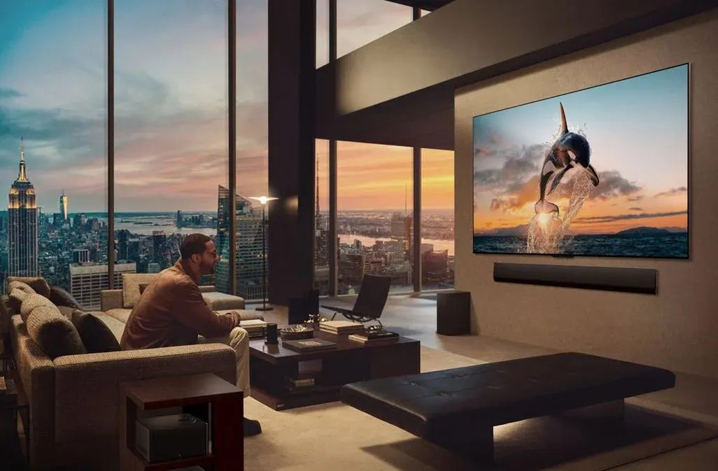 Ein Mann auf einem Sofa schaut einen TV mit einem springenden Orca. Im Hintergrund ist die New Yorker Skyline mit dem Empire State Building bei Sonnenuntergang zu sehen.