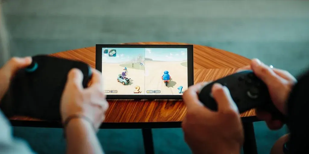 Zwei Personen spielen mit schwarzen Controllern ein Rennspiel auf einem geteilten Bildschirm. Bunte Charaktere fahren auf einer Sandstrecke. Das Display liegt auf einem Holztisch.