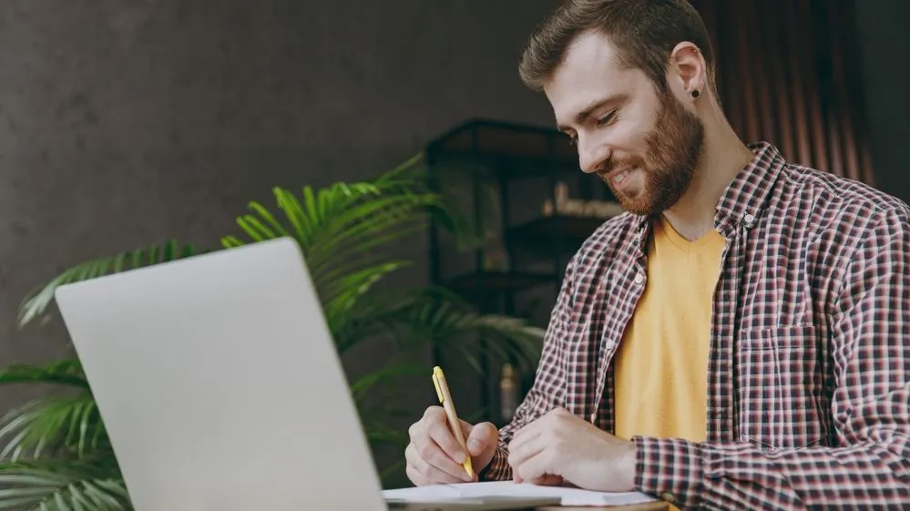 Mann mit Bart schreibt Notizen während er am Laptop arbeitet