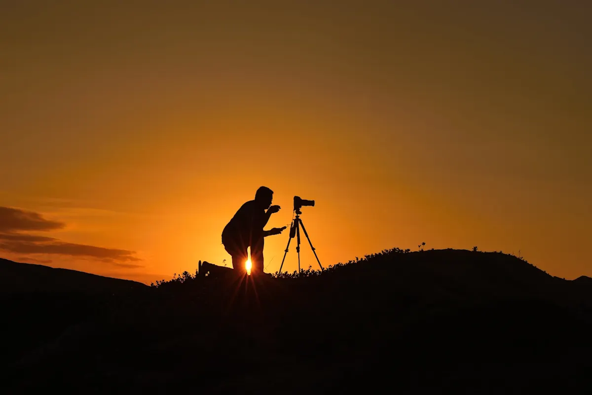 : Silhouette eines Fotografen, der vor einer Kamera auf einem Stativ kniet. Dahinter leuchtet ein durch die untergehende Sonne oranger Himmel.