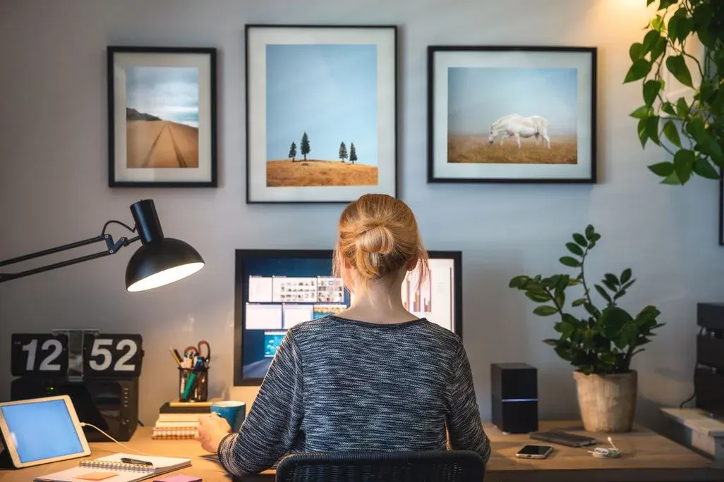 Person am Schreibtisch vor Monitor, mit Lampe, Tablet, Notizbuch. An der Wand drei gerahmte Bilder: Strand, Bäume, Pferde; grüne Pflanzen.