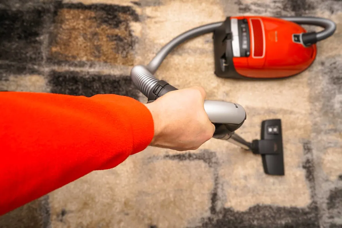 Ein Arm mit rotem Pullover hält den Griff eines roten Staubsaugers, der auf einem braungemusterten Teppich steht.