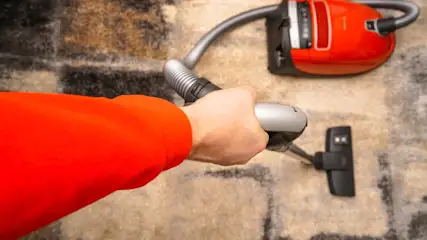 Ein Arm mit rotem Pullover hält den Griff eines roten Staubsaugers, der auf einem braungemusterten Teppich steht.