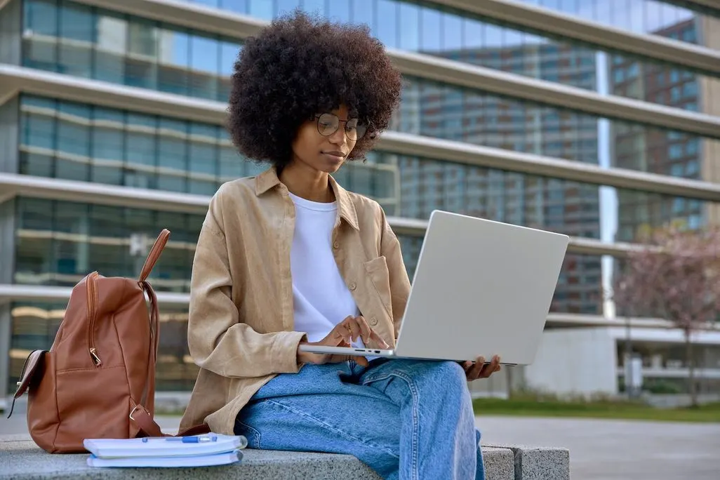 Eine junge Frau nutzt ihren Laptop im Freien vor einem Bürogebäude.