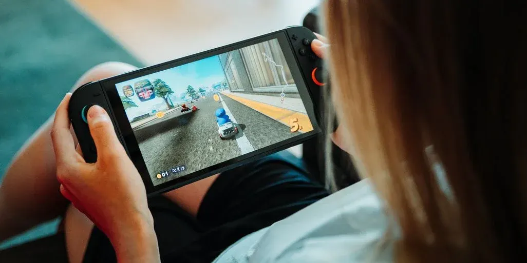 Hände halten eine schwarze Handheld-Konsole, auf deren Bildschirm ein Rennspiel mit einem blauen Kart auf einer Straße mit gelbem Streifen zu sehen ist.