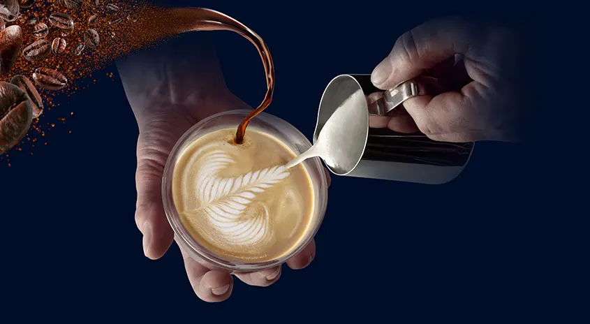 Eine Hand hält eine Tasse Kaffee, während eine andere Milch aus einem silbernen Krug einschenkt. Kaffeebohnen links.